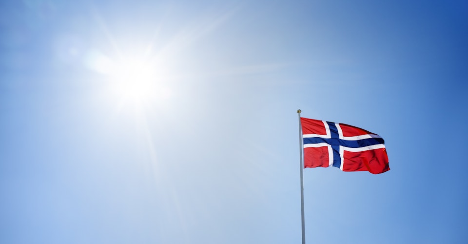 Det norske flagget på toppen av en flaggstang. Blå himmel med sol i bakgrunnen. Foto.
