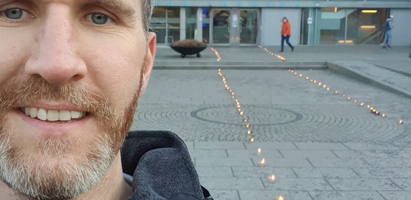 Mairus Nilsen, selfie foran bygg med fakler i forkant. Foto.