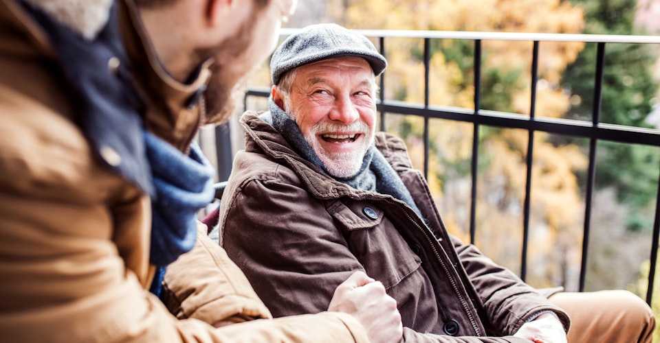 Eldre mann i ytterjakke og sixpence, smiler på en balkong. Foto.