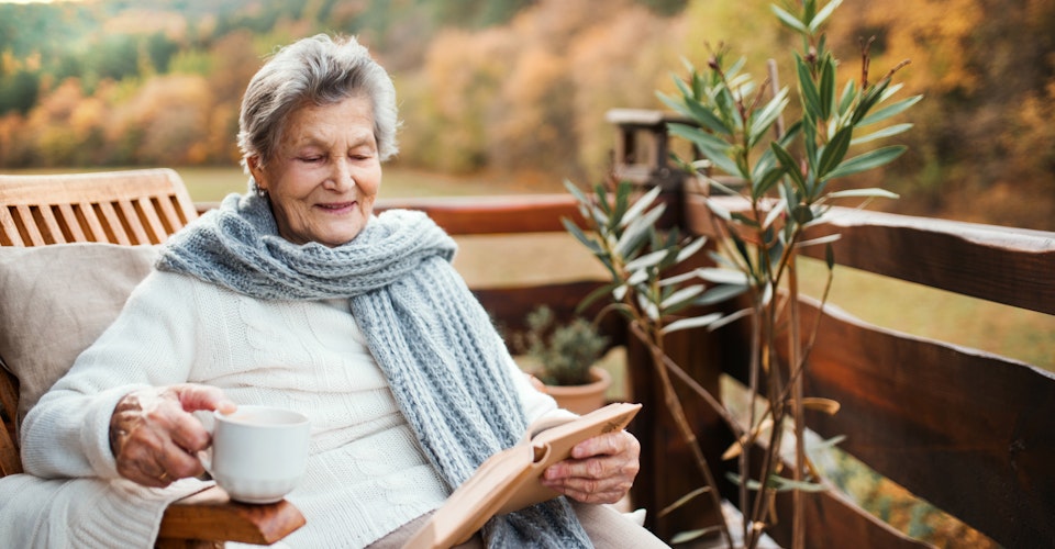 Eldre kvinne sitter i en stol utenfor og leser i en bok. Kaffekopp i en hånd. I bakgrunn trær i høstfarger. Foto.