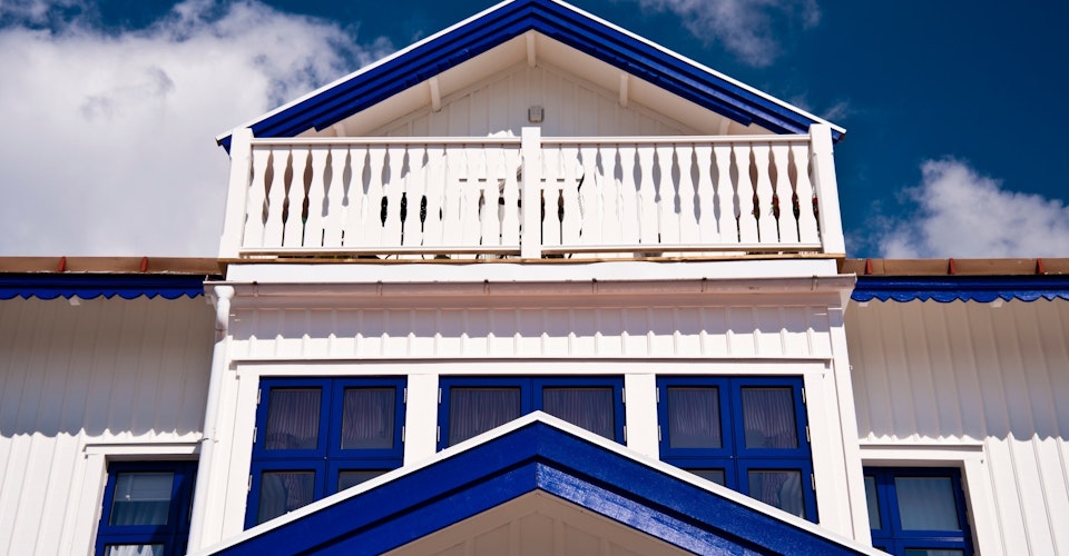 Stort hvitt hus med blå lister og møner, sett nedenfra mot blå himmel. Foto.